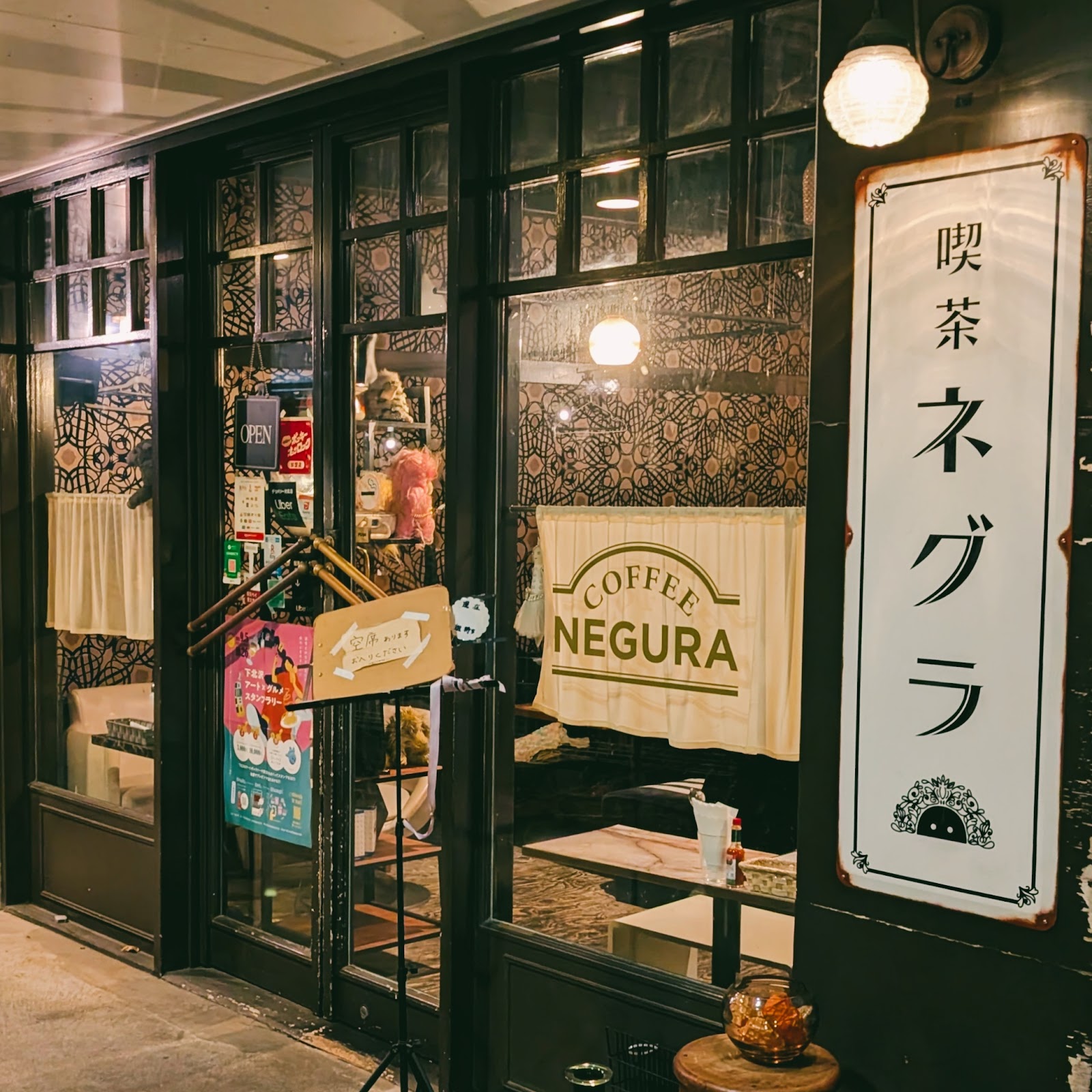 夜の外観、黒いサッシの格子窓にボタニカル柄の壁紙が透け、「喫茶ネグラ」縦看板と丸い白いペンダントライトが灯る