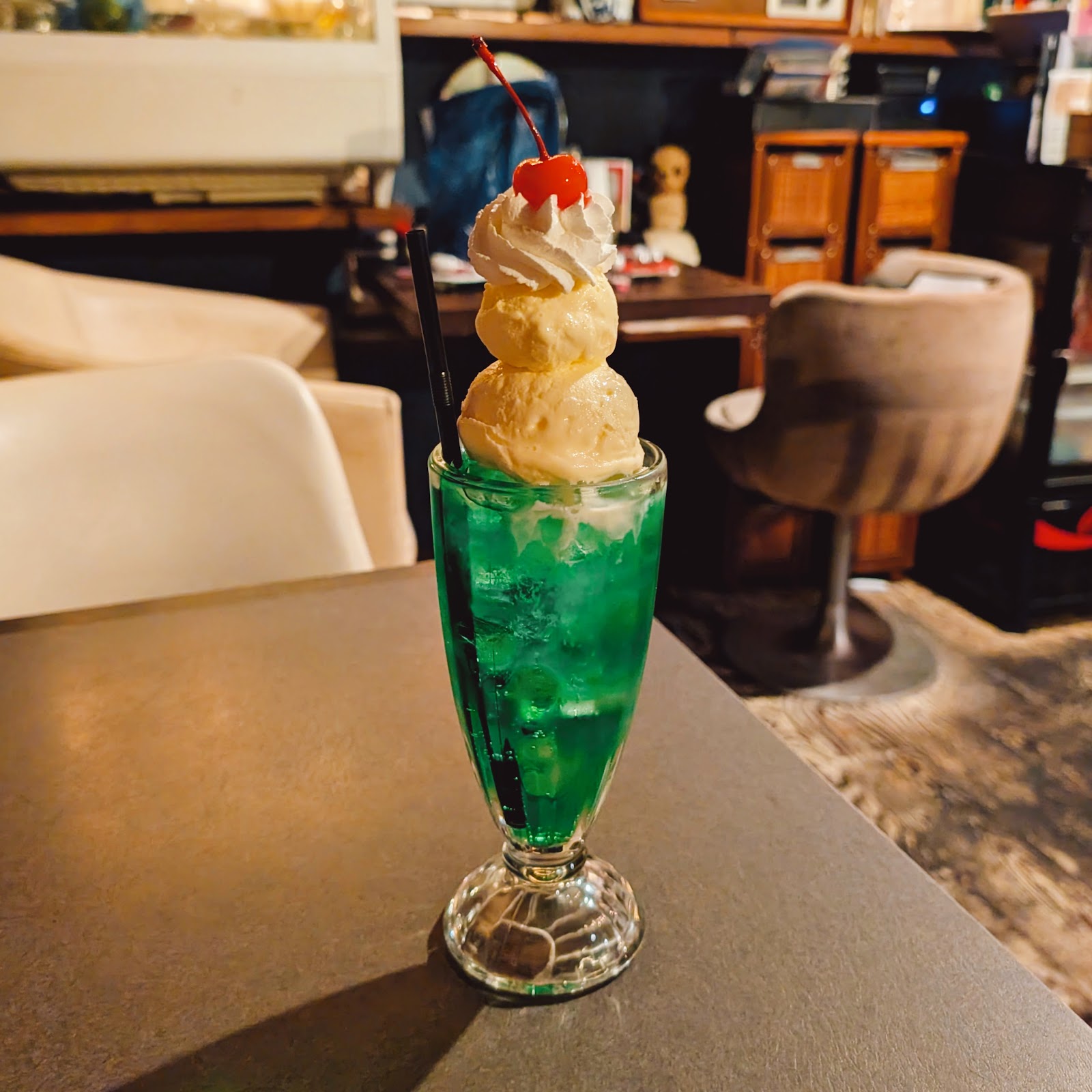鮮やかな緑色のメロンクリームソーダ、バニラアイス2玉とホイップクリームと赤いチェリーが乗ったグラスが手前に置かれ、奥に薄暗い昭和レトロな店内の棚と丸椅子が見える