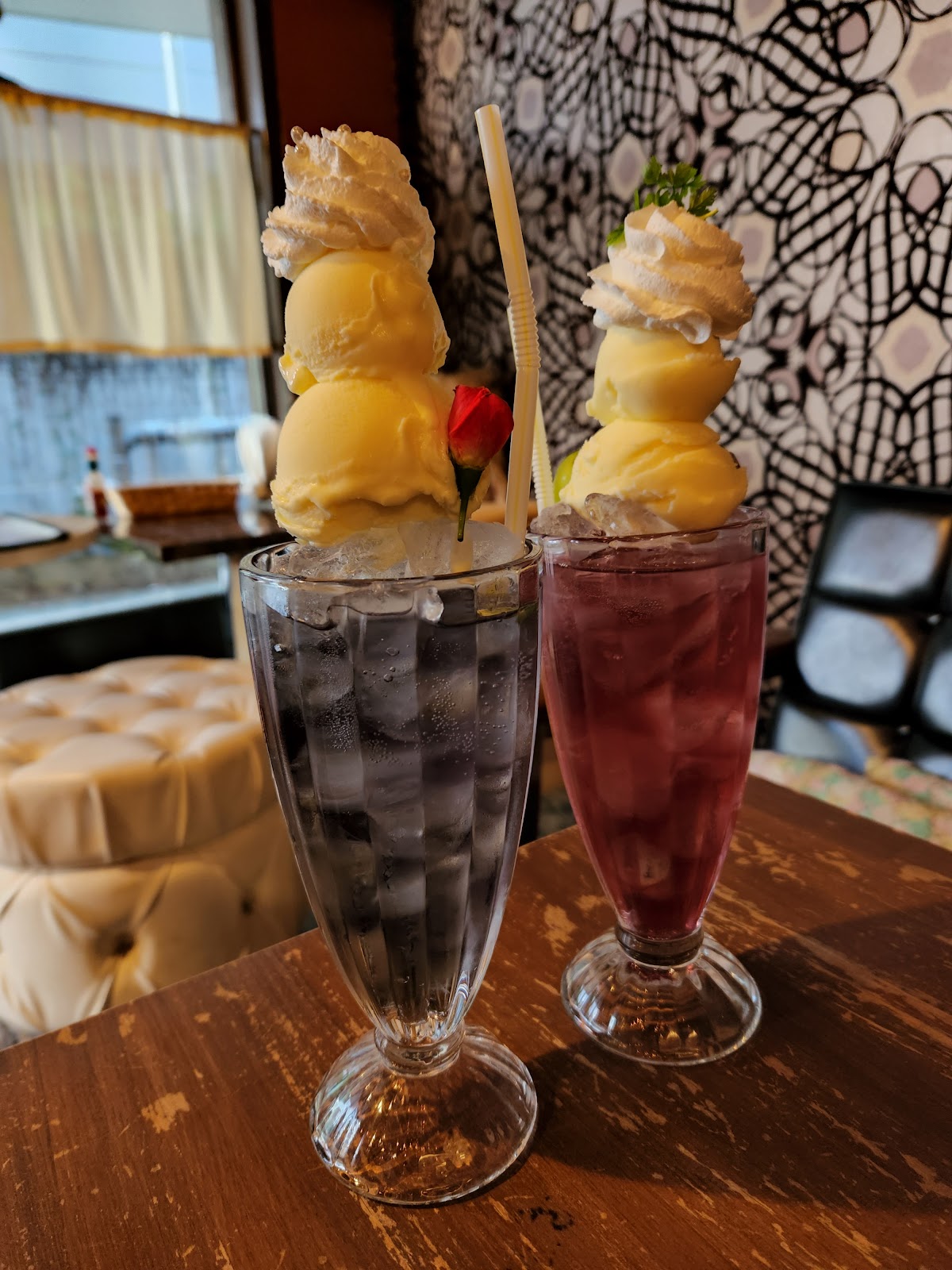 木製テーブルの上に並ぶ黒と紫2色のクリームソーダ、バニラアイスを高く積み上げホイップクリームをのせ、背景にボタニカル柄壁紙とソファー席が見える